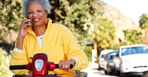 Elderly woman in yellow jersey smiling and riding mobility scooter down the street