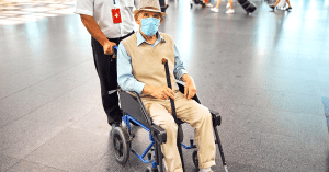 Elderly man with face mask and cane sitting in wheelchair at airport, being assisted by attendant