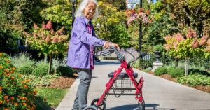 Elderly woman walking in park with red four wheeled walker with seat